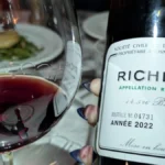 A dark red wine glass with swirling liquid under warm lighting next to wooden barrels.