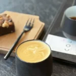 A steaming pour-over coffee in a glass vessel on a rustic wooden table with sunlight filtering through a green curtain.