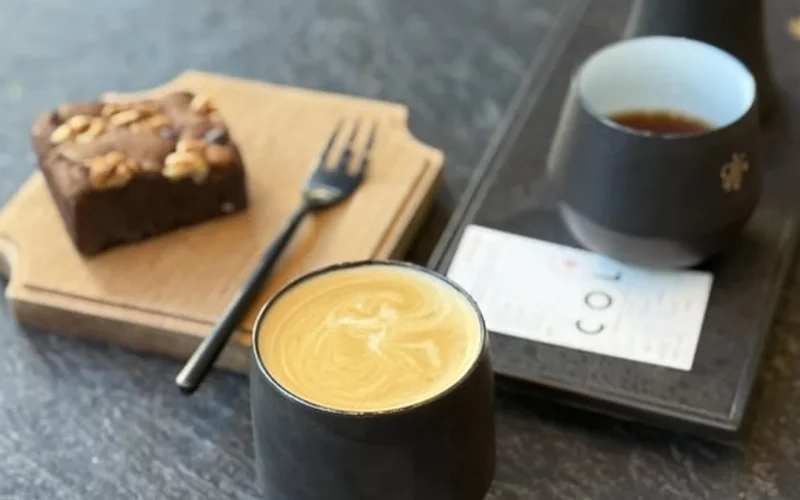 A steaming pour-over coffee in a glass vessel on a rustic wooden table with sunlight filtering through a green curtain.
