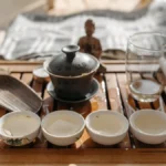 Gaiwan with Longjing Dragonwell tea, steam rising, wooden tray and bamboo strainer in soft morning light.