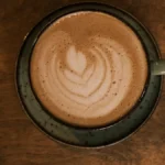 Golden espresso foam pouring into a copper cup with steam rising and coffee beans in the background.