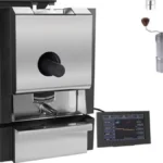 Black coffee roaster emitting steam in rustic kitchen setting with coffee beans and parchment nearby.