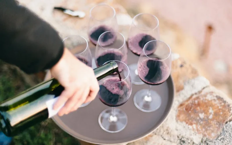 A dusty red wine bottle pouring into a dark glass with a wooden table and flickering candlelight.