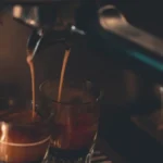 Espresso being poured from Bambino Plus into a dark ceramic cup with steam rising and warm light from a window.