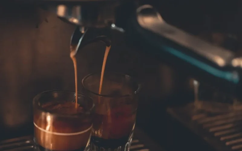 Espresso being poured from Bambino Plus into a dark ceramic cup with steam rising and warm light from a window.