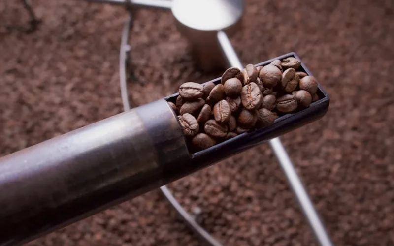Dark coffee beans spilling from roaster drum with steam and golden light casting shadows on wooden floor.