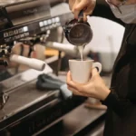 Barista pours oat milk into latte art cup with steam rising in bright morning light.