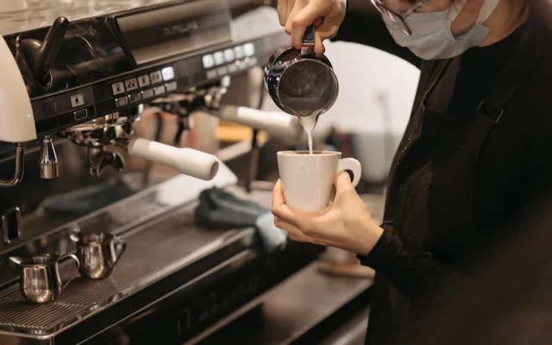 Barista pours oat milk into latte art cup with steam rising in bright morning light.