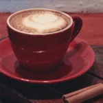 A steaming cortado coffee in a porcelain mug on a white counter with espresso machine and coffee bean.