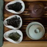 A steaming gaiwan with tea liquor and dried leaves on a wooden tray with bamboo tools under warm light.