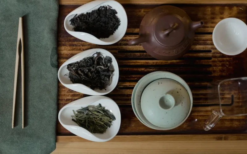 A steaming gaiwan with tea liquor and dried leaves on a wooden tray with bamboo tools under warm light.
