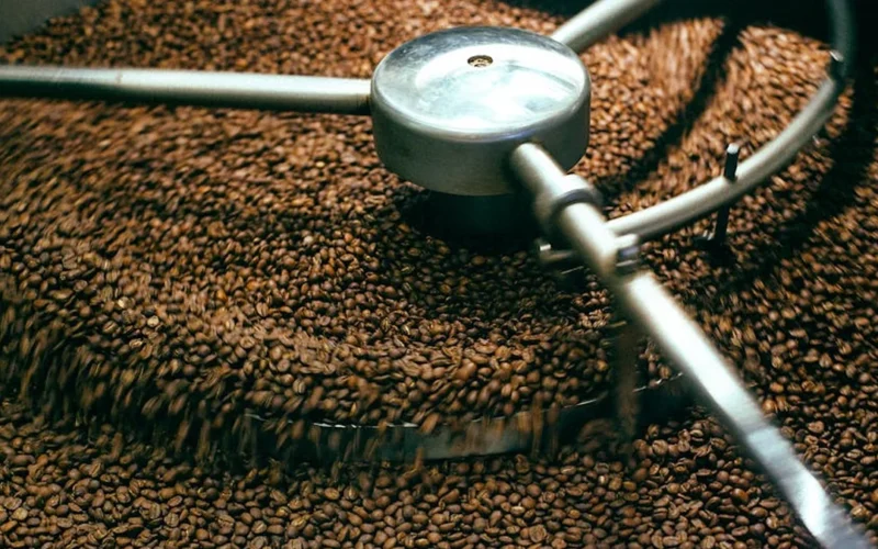 Dark roasted coffee beans steaming near a roasting machine under warm lighting.