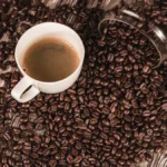 Coffee beans spilling from a bag onto a wooden table with a steaming mug nearby.