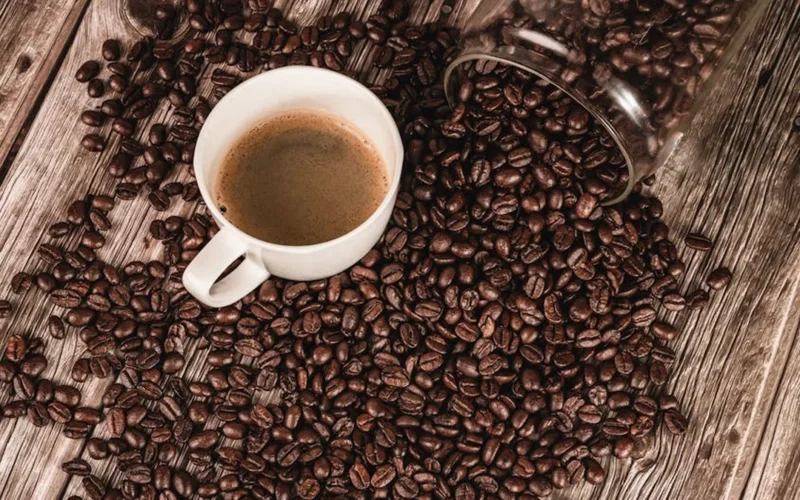 Coffee beans spilling from a bag onto a wooden table with a steaming mug nearby.