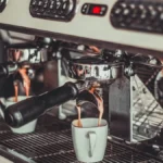 Dark coffee beans grinding into portafilter with espresso steaming from white cup
