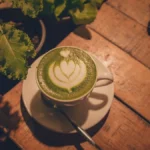 A wooden table displays a steaming hojicha mug and a matcha latte under warm golden light.