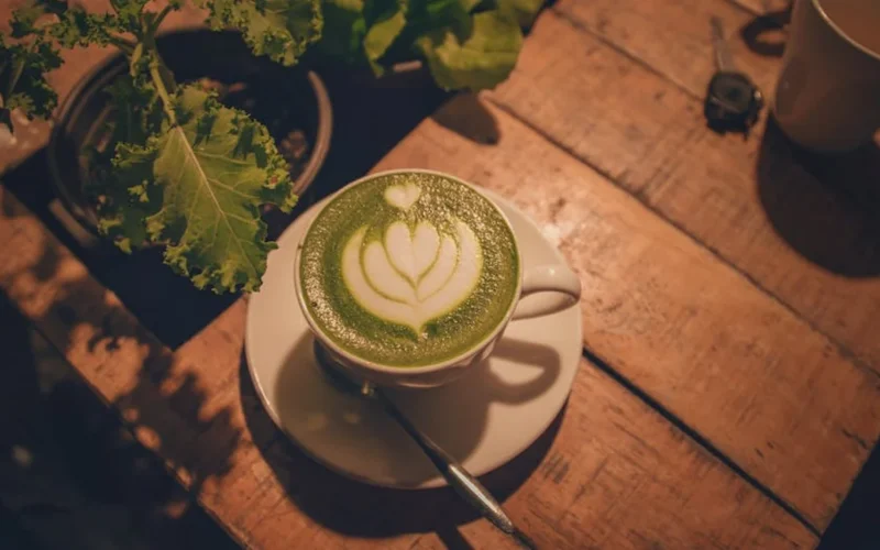 A wooden table displays a steaming hojicha mug and a matcha latte under warm golden light.
