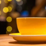 A dark roast coffee cup with beans on a wooden table next to a vintage grinder under warm lighting.