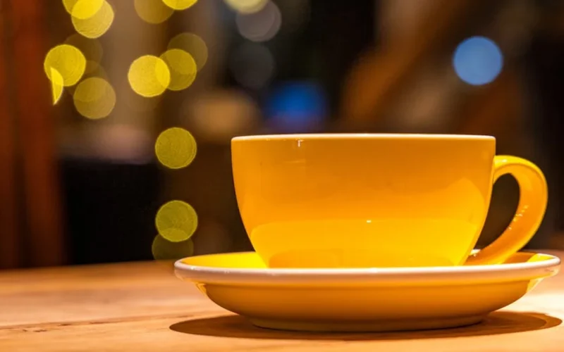 A dark roast coffee cup with beans on a wooden table next to a vintage grinder under warm lighting.