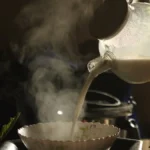 Steam rising from a glass of water over a steaming coffee cup with a metallic brewing device in the background.