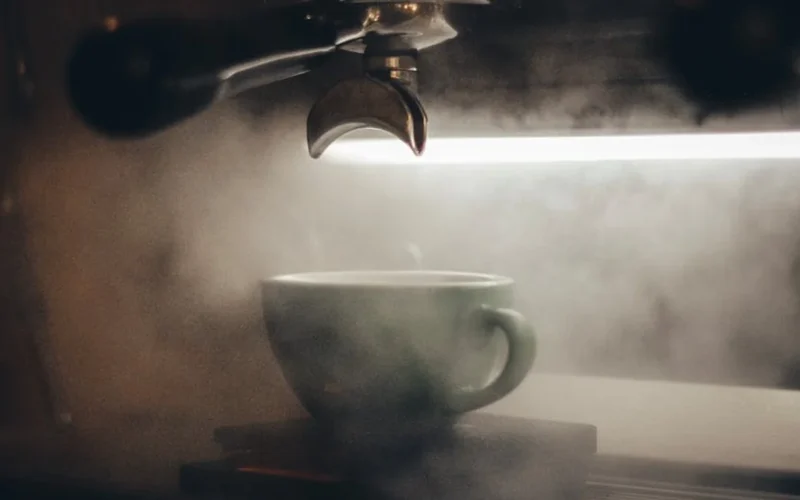 Brass coffee machine steaming milk into artistic froth on marble counter under warm lighting.
