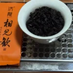 Ajar of dark, wiry tea leaves with charcoal roast aroma under golden light on a wooden table.