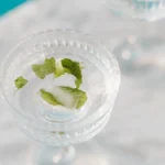 A glass jar with crushed mint and basil steeping in iced tea, surrounded by fresh herbs and a ceramic pitcher.