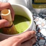 A steaming matcha latte in a ceramic cup on a wooden counter with traditional tea ceremony elements nearby.