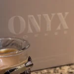 Dark roast coffee beans in a ceramic jar with steam rising from a pour-over coffee setup under warm lighting.