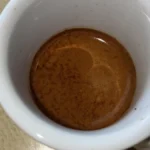 Dark roast coffee beans spilling into black cup with steam from espresso machine