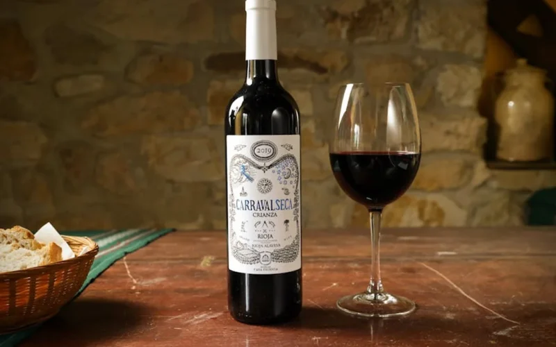 A dark wooden table features a Cabernet Franc wine bottle, glass of wine, and open notebook under warm lighting.