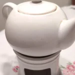 Porcelain teapot with wide spout pouring loose leaf tea into a white ceramic mug under soft natural light.