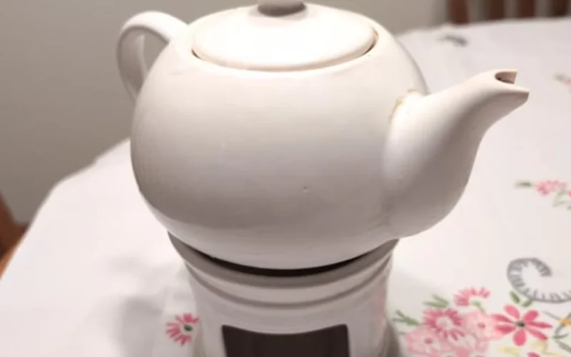 Porcelain teapot with wide spout pouring loose leaf tea into a white ceramic mug under soft natural light.