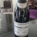 Dusty Romanée-Conti bottle with intact cork on dark wooden table under warm lighting