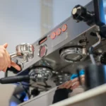 Two high-end espresso machines side by side in a modern coffee bar with steam rising and light reflecting off their surf