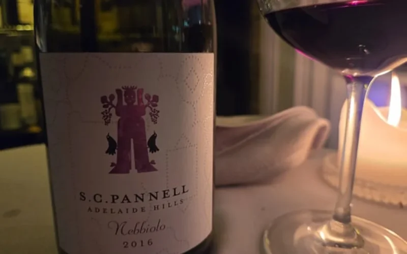 A deep red Nebbiolo wine in a decanter against aged oak barrels in a rustic cellar.
