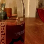 A dark red wine glass with swirling liquid sits on a rustic wooden table surrounded by dried herbs under golden lighting