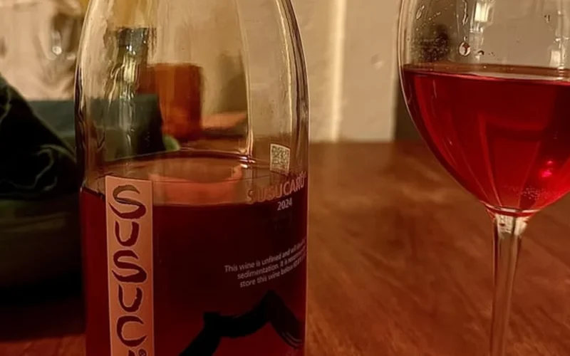 A dark red wine glass with swirling liquid sits on a rustic wooden table surrounded by dried herbs under golden lighting