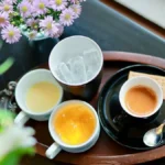 A steaming tea cup on a bamboo tray with a kettle and sunlight casting shadows on a wooden table.