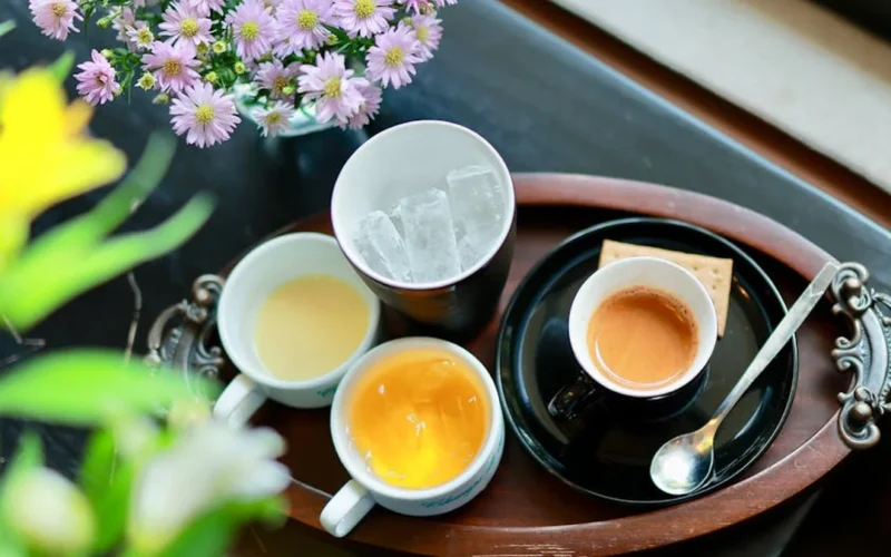 A steaming tea cup on a bamboo tray with a kettle and sunlight casting shadows on a wooden table.