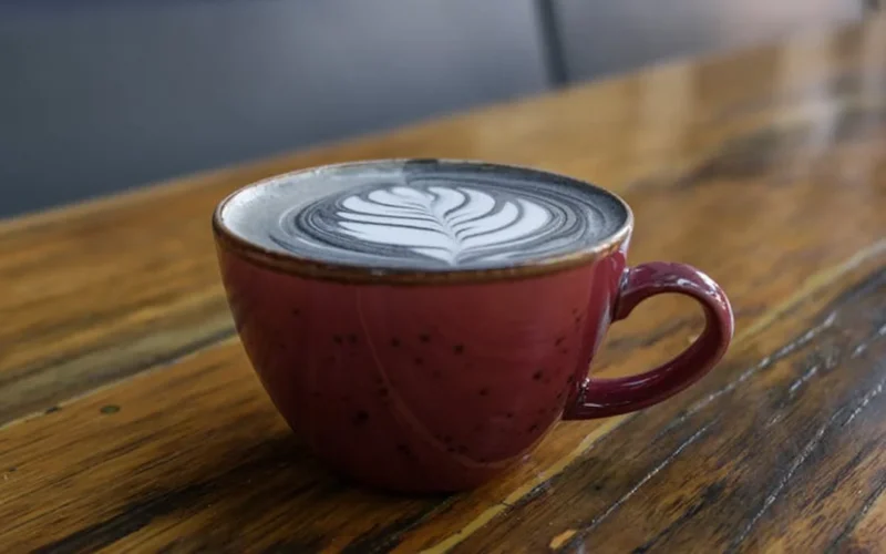 A steaming coffee mug on a wooden table under a sunrise with coffee beans and a clock nearby.