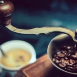 Black espresso grinder grinding beans with golden light reflecting off its surface next to a steaming espresso shot.
