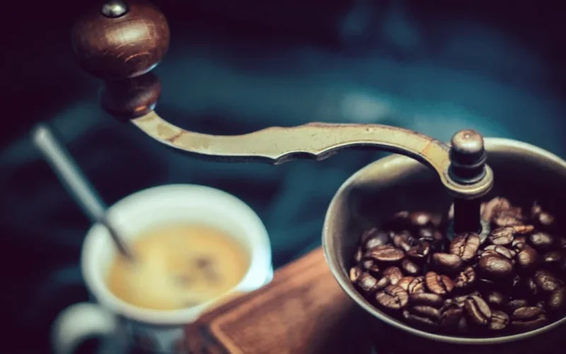 Black espresso grinder grinding beans with golden light reflecting off its surface next to a steaming espresso shot.