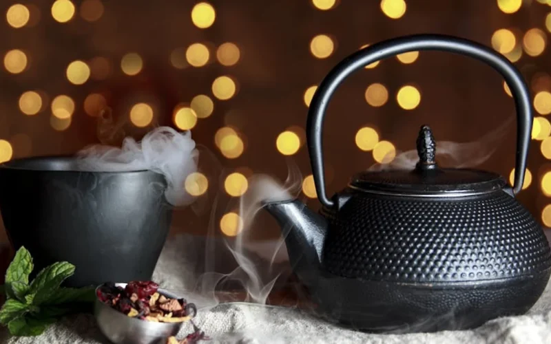 A glass teapot filled with shimmering black tea leaves and rising steam under warm lighting.