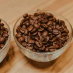 Coffee beans spilling from a bag onto a wooden table with steam rising from a French press under warm light.