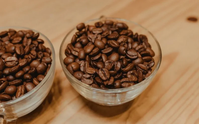 Coffee beans spilling from a bag onto a wooden table with steam rising from a French press under warm light.