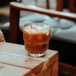 A glass jar filled with dark coffee and ice cubes under warm lighting on a wooden table.