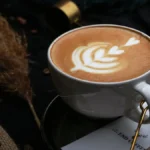 A coffee cup with latte art and coffee beans on a wooden surface next to a scale and notebook.