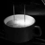 Dark espresso dripping into black ceramic cup with steam and shadowy lighting.