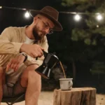 Group of professionals gathering around a campfire with coffee mugs in a wooded setting.